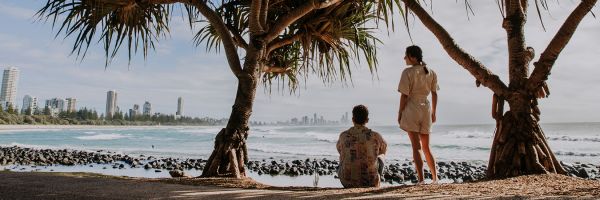 Couple shaded by large beach trees sitting on the beach looking out at skyscrapers on the coastline